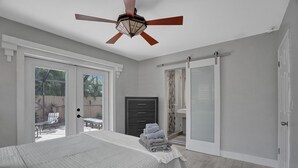 A bedroom with a bed, a ceiling fan, a sliding glass door leading to a patio, a dark-colored dresser, and a shower area. - Beautiful Home; Intimate Backyard with Private Pool Sleeps 8! (Wilton Manors)