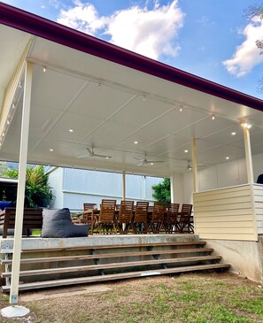 Eine überdachte Terrasse im Freien mit einer Betontreppe, die zu einer Sitzgruppe mit Holzstühlen und einer Bank führt.