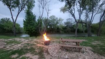 A campfire with a picnic table and a stone fire pit in a grassy area with trees and a river in the background.
