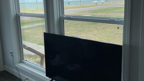 Une chambre dotée d’une grande fenêtre donnant sur une plage, d’un téléviseur à écran plat posé sur un meuble blanc et d’une prise de courant au mur.