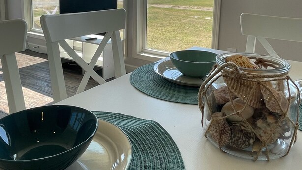Un coin repas avec une table dressée pour deux personnes, de grandes fenêtres offrant une vue sur un pont et un espace verdoyant, et un vase décoratif rempli de coquillages posé sur la table.