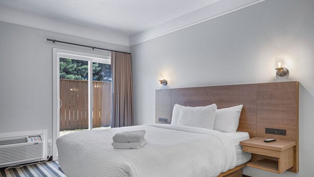 A hotel room with a large bed, a bedside table with a remote control, a wall-mounted light, and a sliding glass door to the outside. - Northwinds | Deluxe King (Canmore)