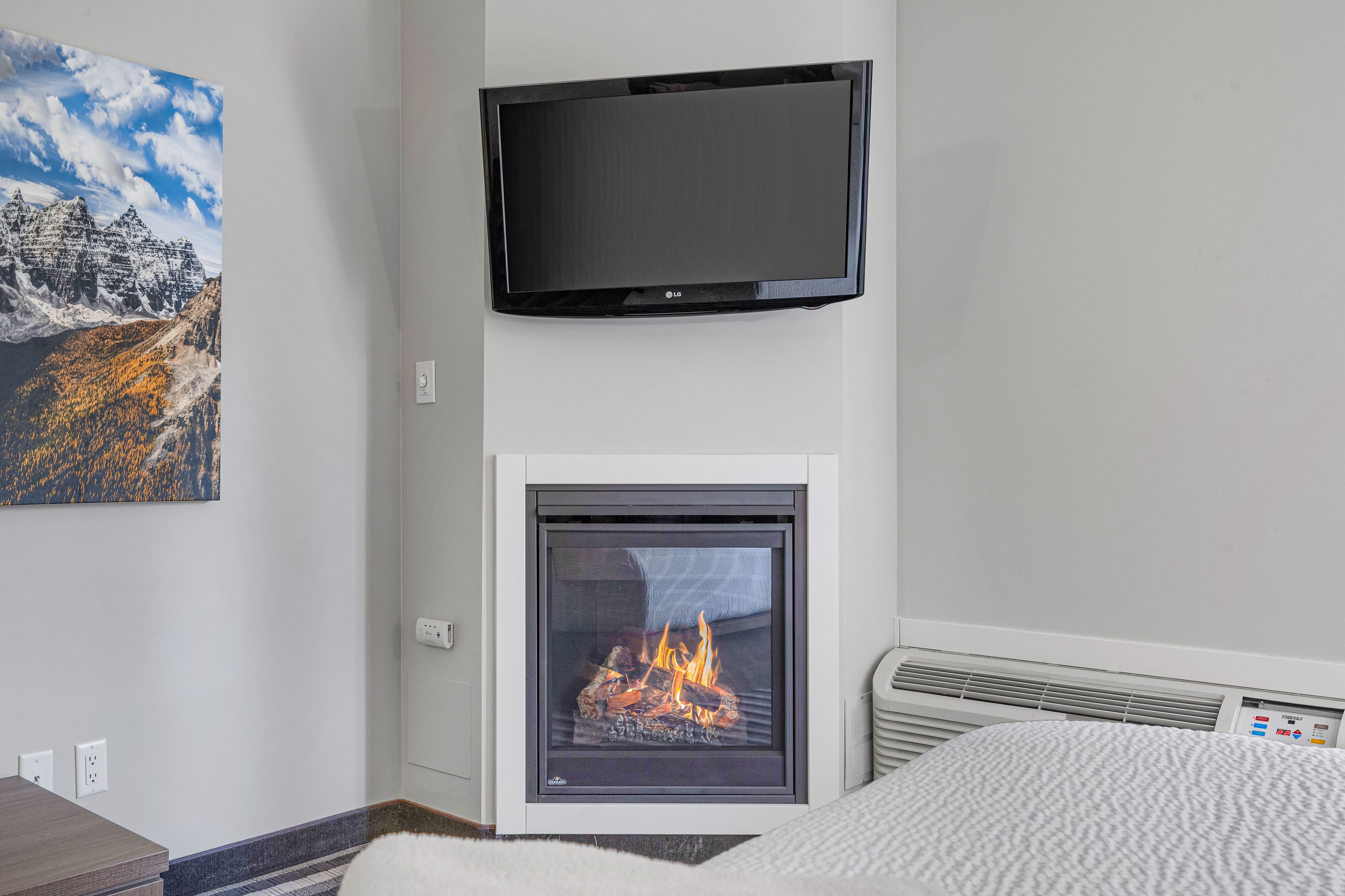 A room with a fireplace, a television mounted on the wall, and a painting of a mountain landscape.