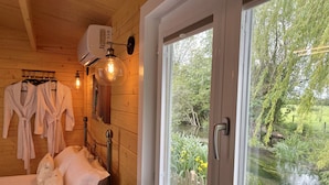 A wooden cabin room with a bed, two robes, a wall-mounted air conditioner, and a window overlooking a river.