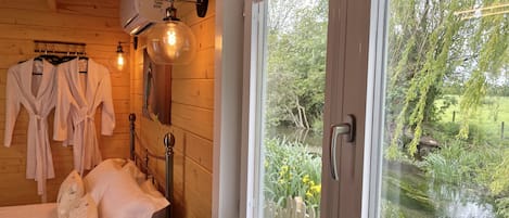 A wooden cabin room with a bed, two robes, a wall-mounted air conditioner, and a window overlooking a river.