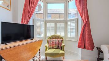 A room with a television on a wooden cabinet, a green armchair with a plaid cushion, and a window with red curtains.