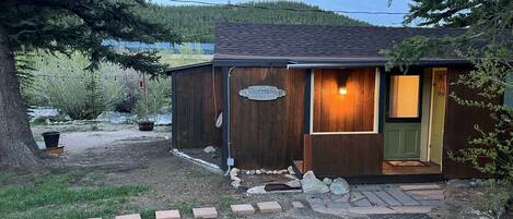 En stuga med en träfasad, en välkomnande skylt, en upplyst entré och en stensatt gångväg som leder fram till dörren.