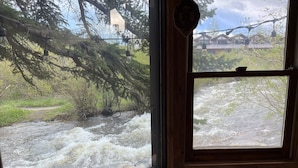 A window view of a fast-flowing river with surrounding greenery and a wooden cabin in the background.