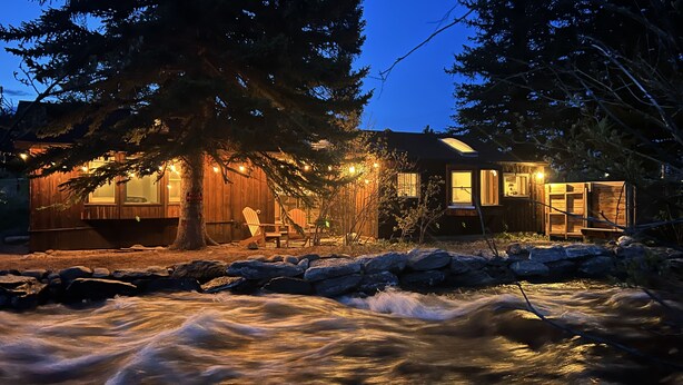 A cabin with a wooden exterior, a large tree in front, and a river in the foreground.
