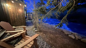 A wooden deck with two Adirondack chairs, a patterned rug, and a small shed with string lights.