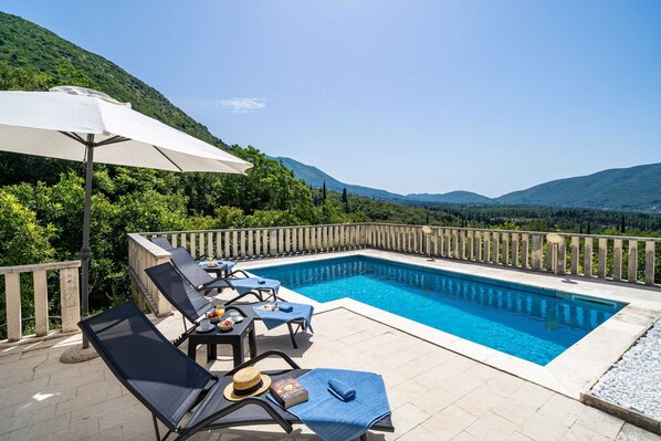 Una piscina con sillas de descanso y sombrillas, rodeada por una terraza de madera y abundante vegetación.