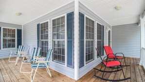 Eine überdachte Veranda mit Holzboden, weißen Wänden und blauen Fensterläden. Vier Schaukelstühle und zwei Lounge-Stühle sind aufgestellt.