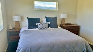 Una cama bien hecha con una colcha gris y dos almohadones decorativos, una mesita de noche con una lámpara y una caja de pañuelos, y un tocador con un pequeño objeto decorativo.