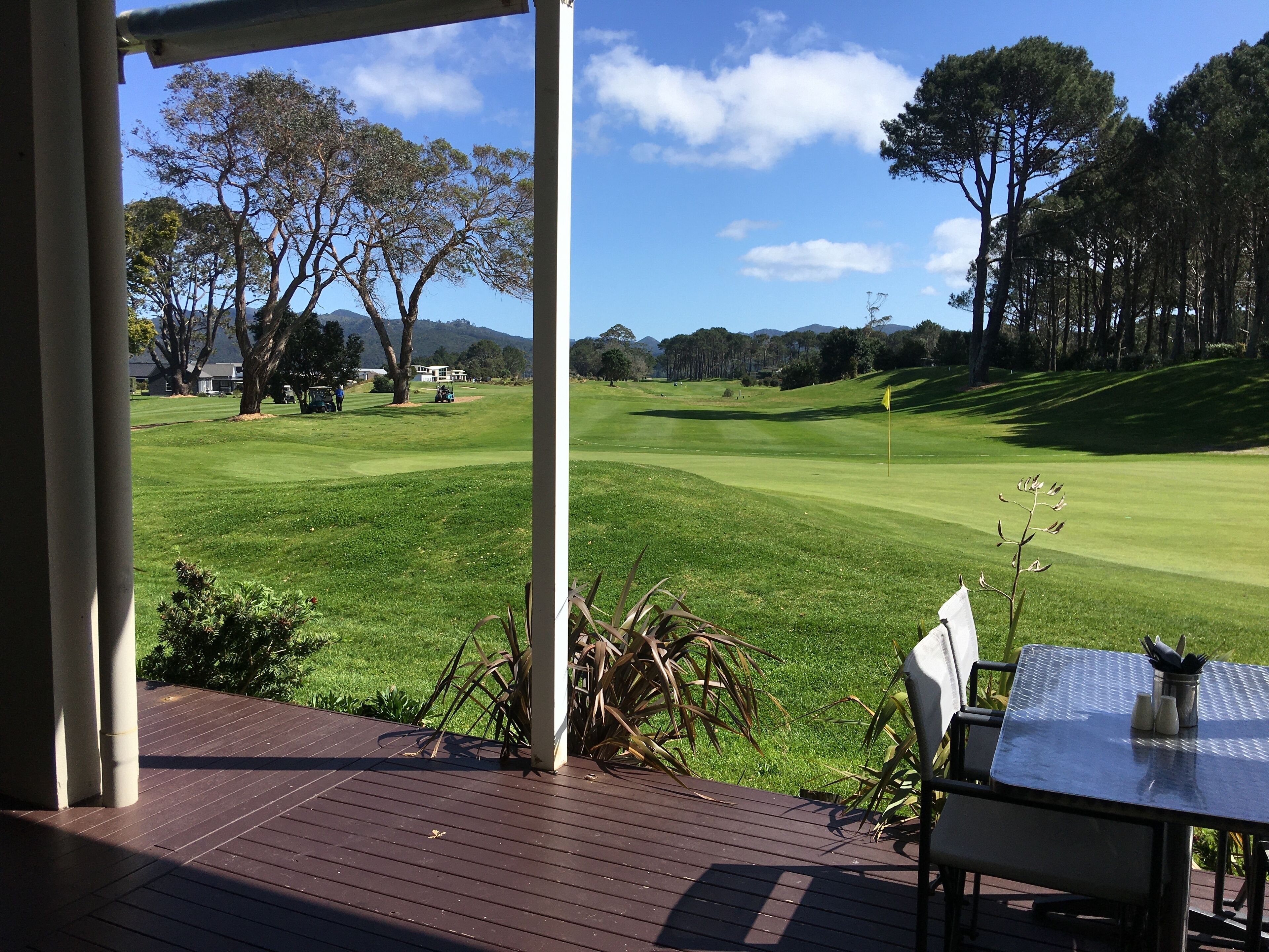 A view from a covered deck overlooking a golf course with a table and chairs, a flag, and distant trees.