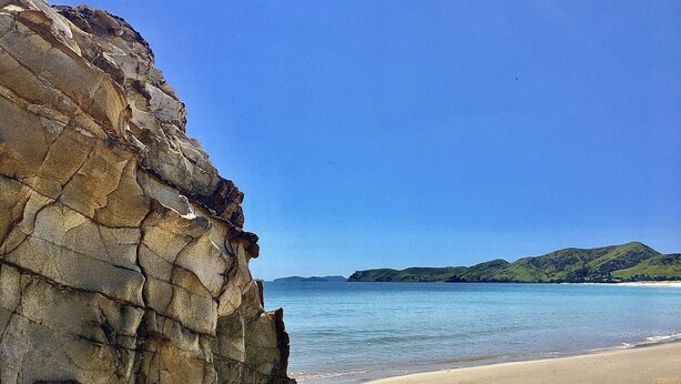 Un paisaje costero con un acantilado rocoso, una playa de arena y colinas verdes distantes bajo un cielo azul despejado.