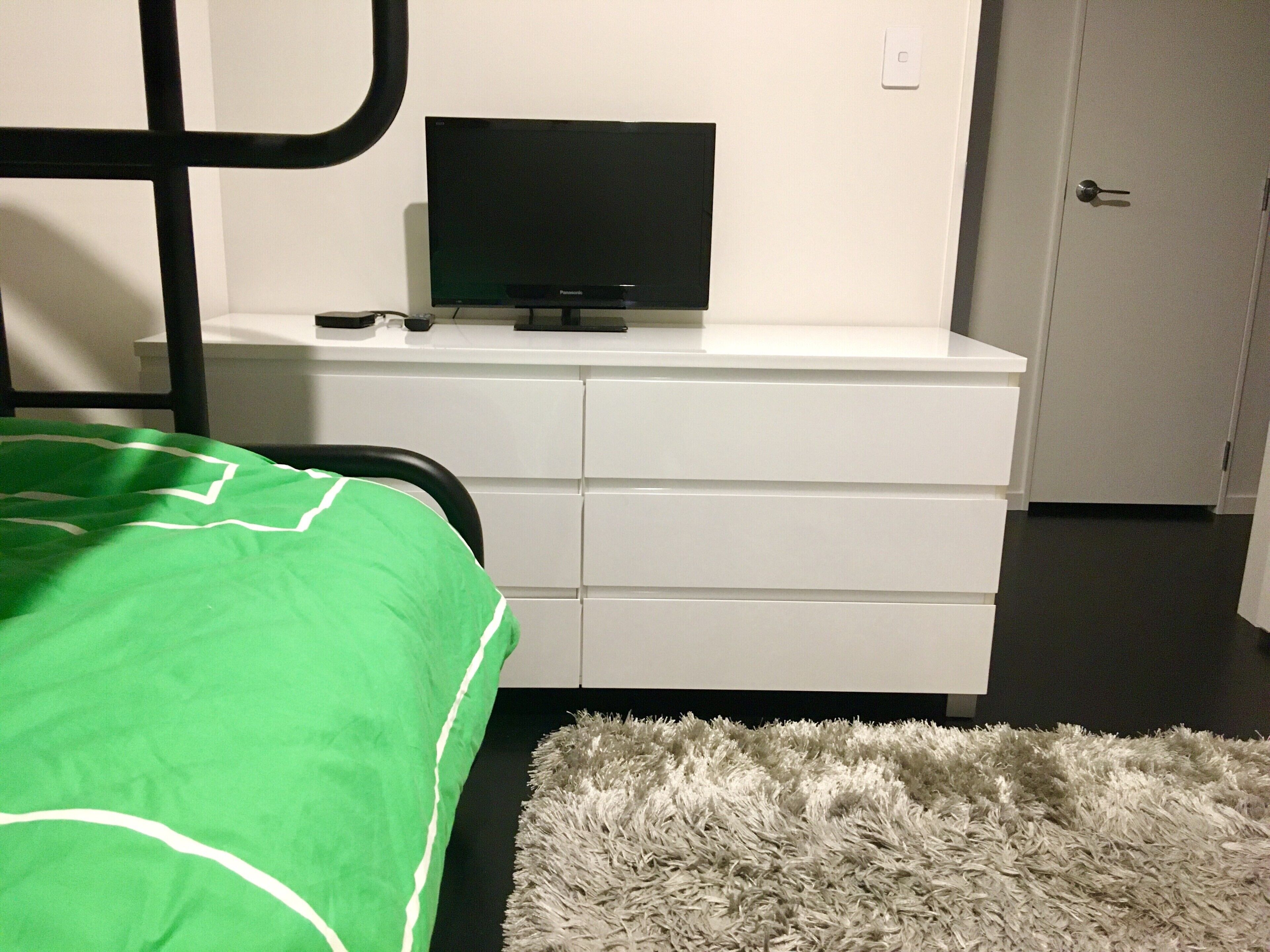 Makuuhuone, jossa on sänky, valkoinen lipasto, jonka päällä on televisio, pehmeä matto ja taustalla ovi.