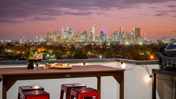 A rooftop terrace with a city skyline view, a table with a bottle, glasses, and a plate of food, and two red stools. - StayCentral Northcote Townhouse With Study (Northcote)