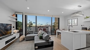 A modern living room with a large TV, grey sofa, and a kitchen area with a white countertop and island. - StayCentral Northcote Townhouse With Study (Northcote)