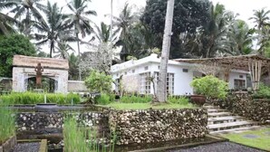 Un edificio tradicional con entrada de piedra, una casa blanca con techo de tejas y un jardín con palmeras y un estanque.