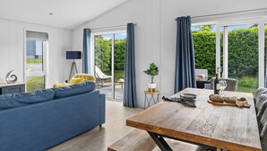 Een moderne woonkamer met een bank, een houten eettafel en uitzicht op een tuin door grote ramen.