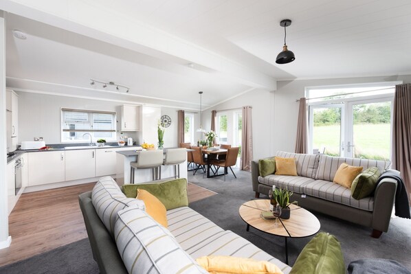 A modern living room with a sofa, a coffee table, and a dining area with a table and chairs. - Daisy Lodge, 6 Roadford Lake Lodges (Lifton)