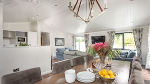 A modern kitchen with built-in appliances, a dining area with a wooden table set for four, and a living room with a large sofa and a TV.