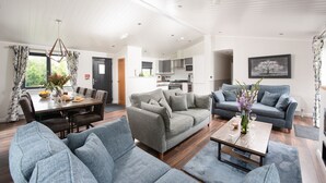 A spacious living room with a dining area, grey sofas, a wooden coffee table, and a kitchen in the background.