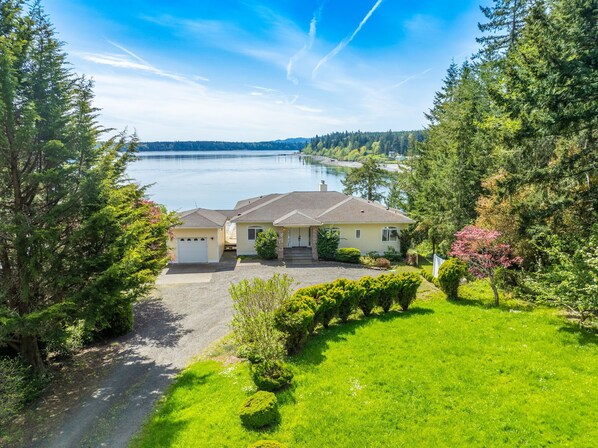 A two-story house with a driveway leading to a garage, surrounded by lush greenery and a lake view.
