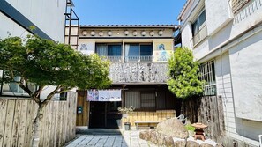 Un edificio japonés tradicional con valla de madera, un pequeño jardín con rocas y un farol de piedra, y un cartel.