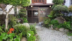 Un edificio japonés tradicional con un letrero de madera, rodeado de abundante vegetación y un camino de grava.