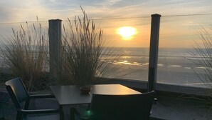 Un área de estar al aire libre con una mesa y sillas, con vista a un cuerpo de agua y un atardecer de fondo.