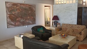 A living room with a large abstract painting, a black leather sofa with a green pillow, a patterned sofa, a coffee table with a bowl, and a wooden cabinet. - Twin Pines Home in Kounoupidiana Chania. (Κουνουπιδιανά)