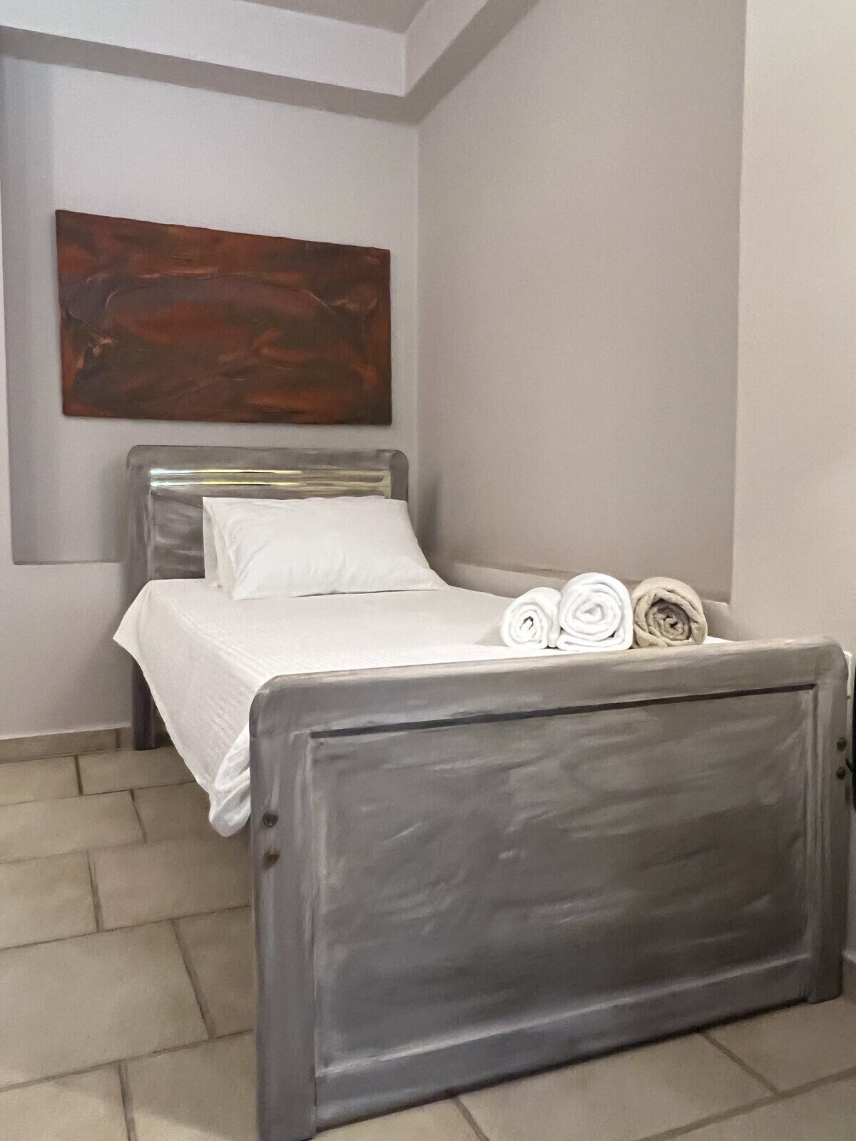 Una cama bien hecha con sábanas blancas y toallas enrolladas, un cabecero gris y un cuadro abstracto grande en la pared.