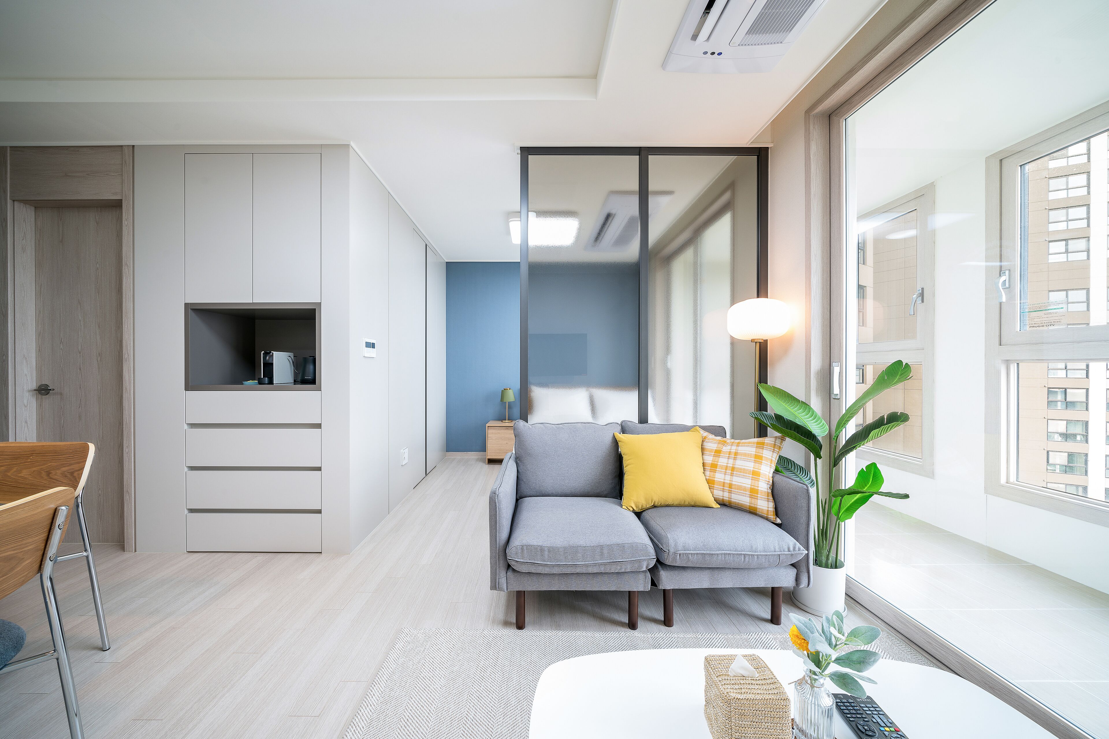 Una sala de estar moderna con un sofá gris, una silla de madera, una mesita con un florero y un amplio ventanal con vistas a la ciudad.