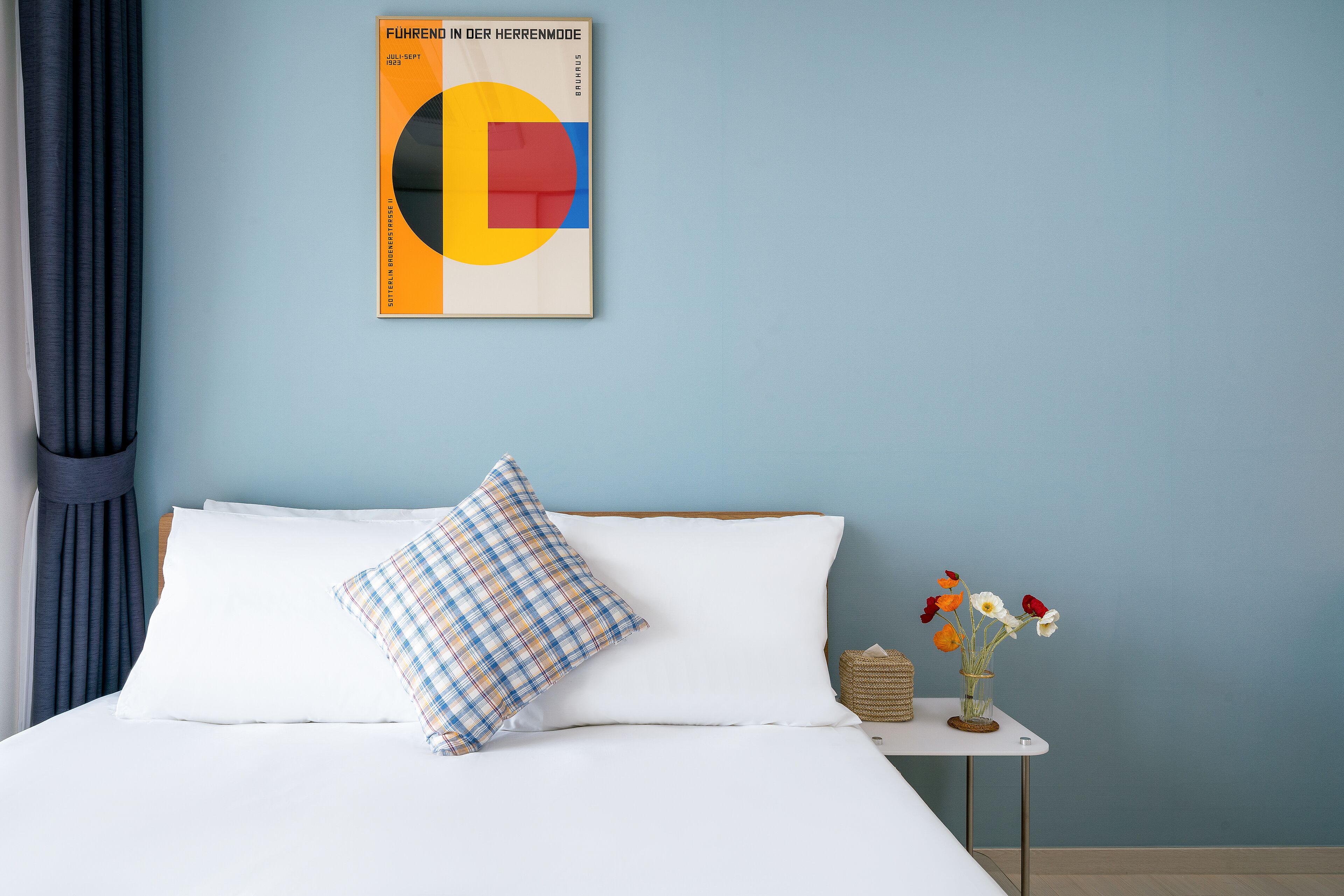 A neatly made bed with white linens, a plaid pillow, and a small bedside table with a vase of flowers.