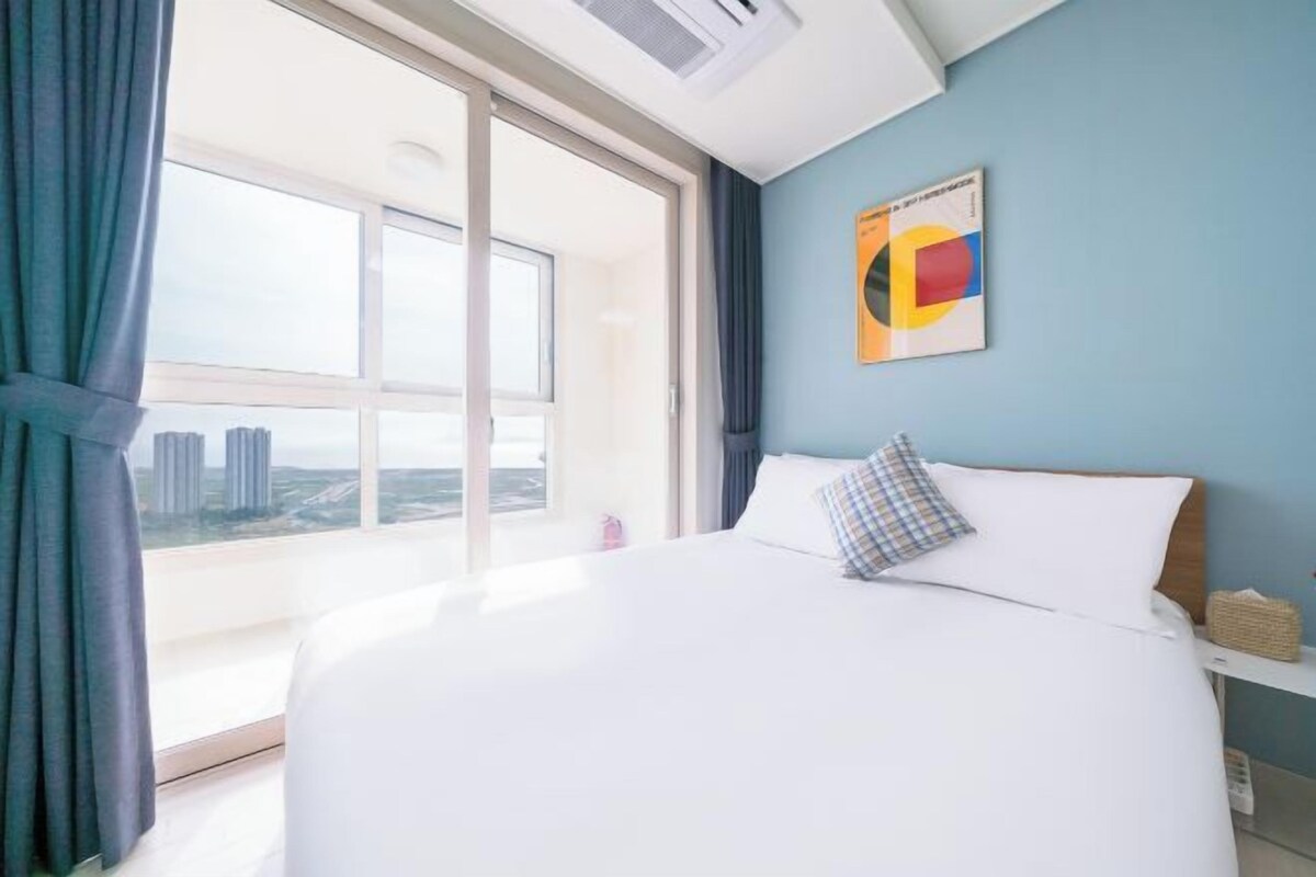 A hotel room with a large bed, a view of the city through a large window, and a wall-mounted artwork.
