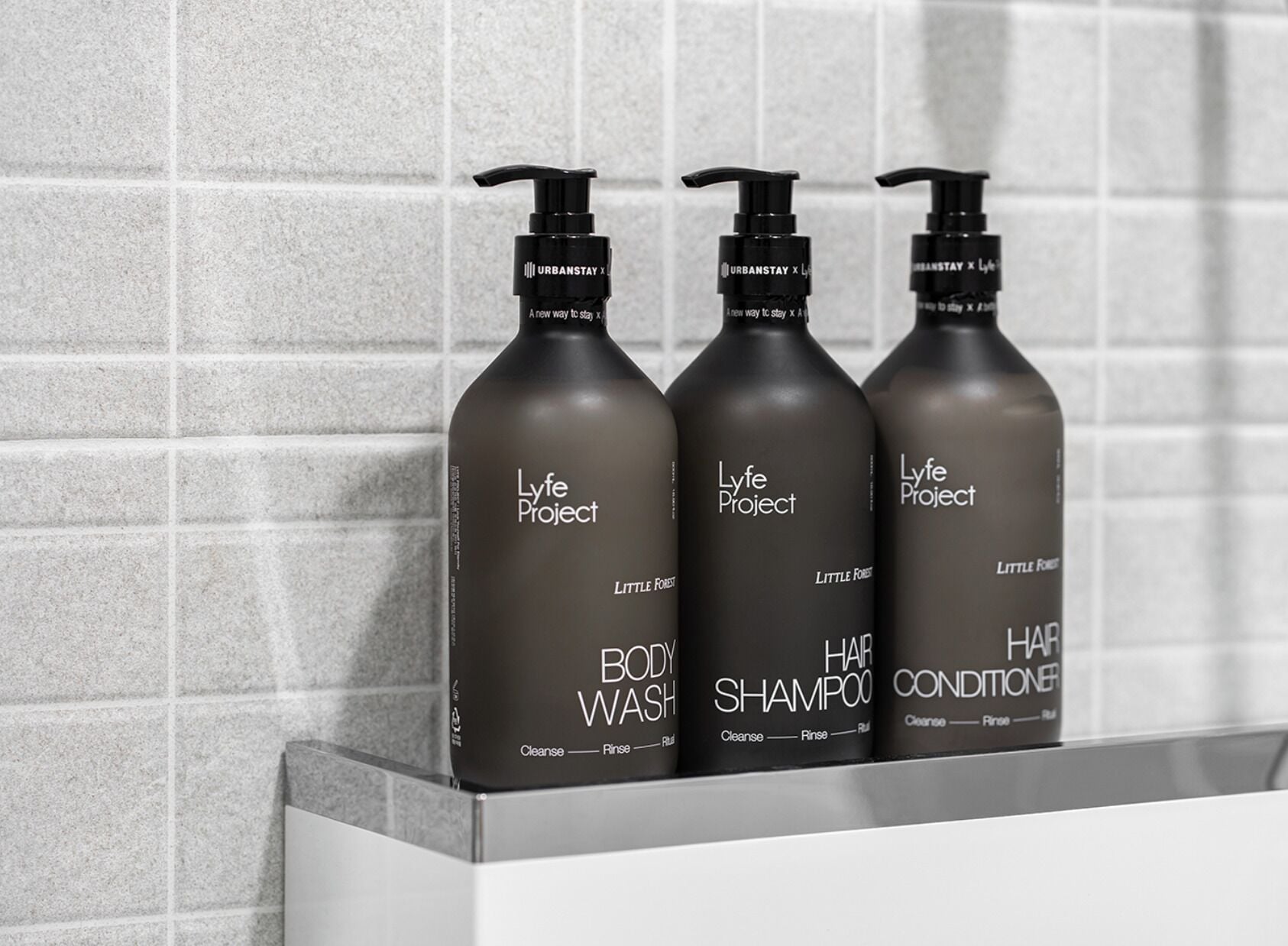 Three bottles of Lyfe Project body wash, shampoo, and conditioner on a shelf against a tiled wall.