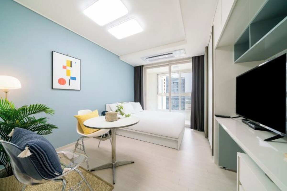 A modern hotel room with a bed, a desk, a chair, a TV, and a window with curtains.