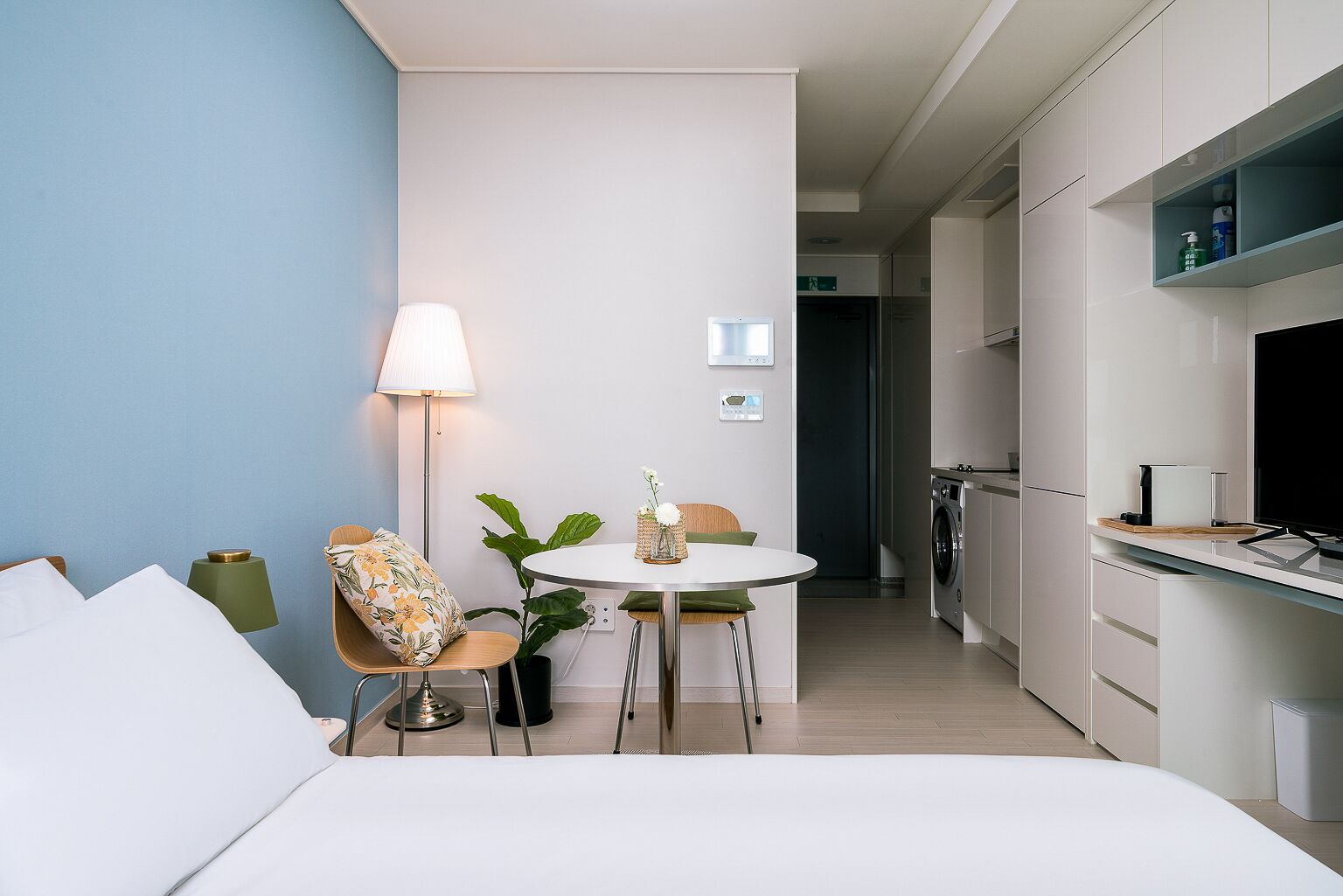Una habitación de hotel moderna con una cama, una mesa redonda con sillas, una lámpara y una planta pequeña. Cuenta con una zona de cocina con lavadora y televisor.