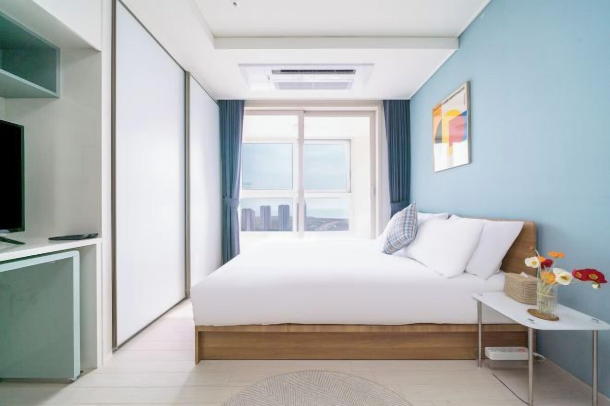 A modern hotel room with a bed, a desk, a TV, and a window with curtains.