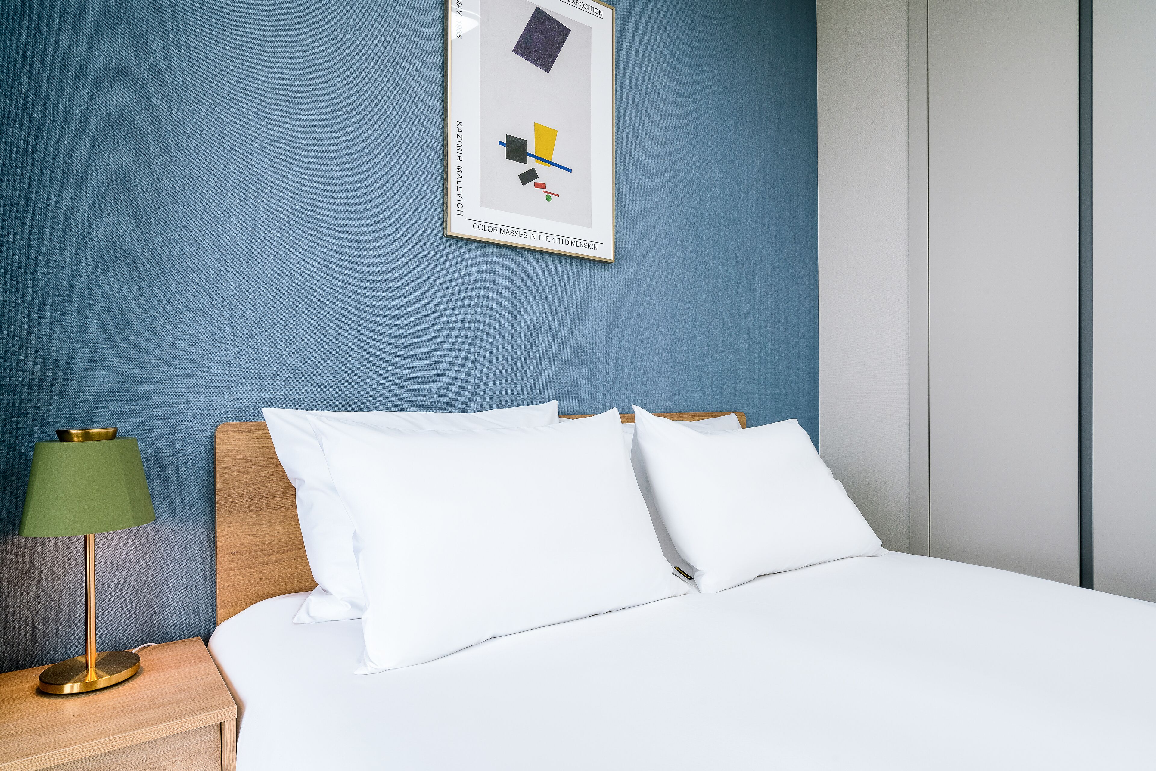 Una cama bien hecha con sábanas blancas, una lámpara verde sobre una mesita de noche de madera y un cuadro abstracto enmarcado en la pared.