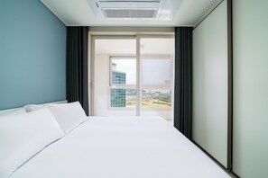 A hotel room with a large bed, a view of a cityscape through a sliding door, and a ceiling air conditioning unit.