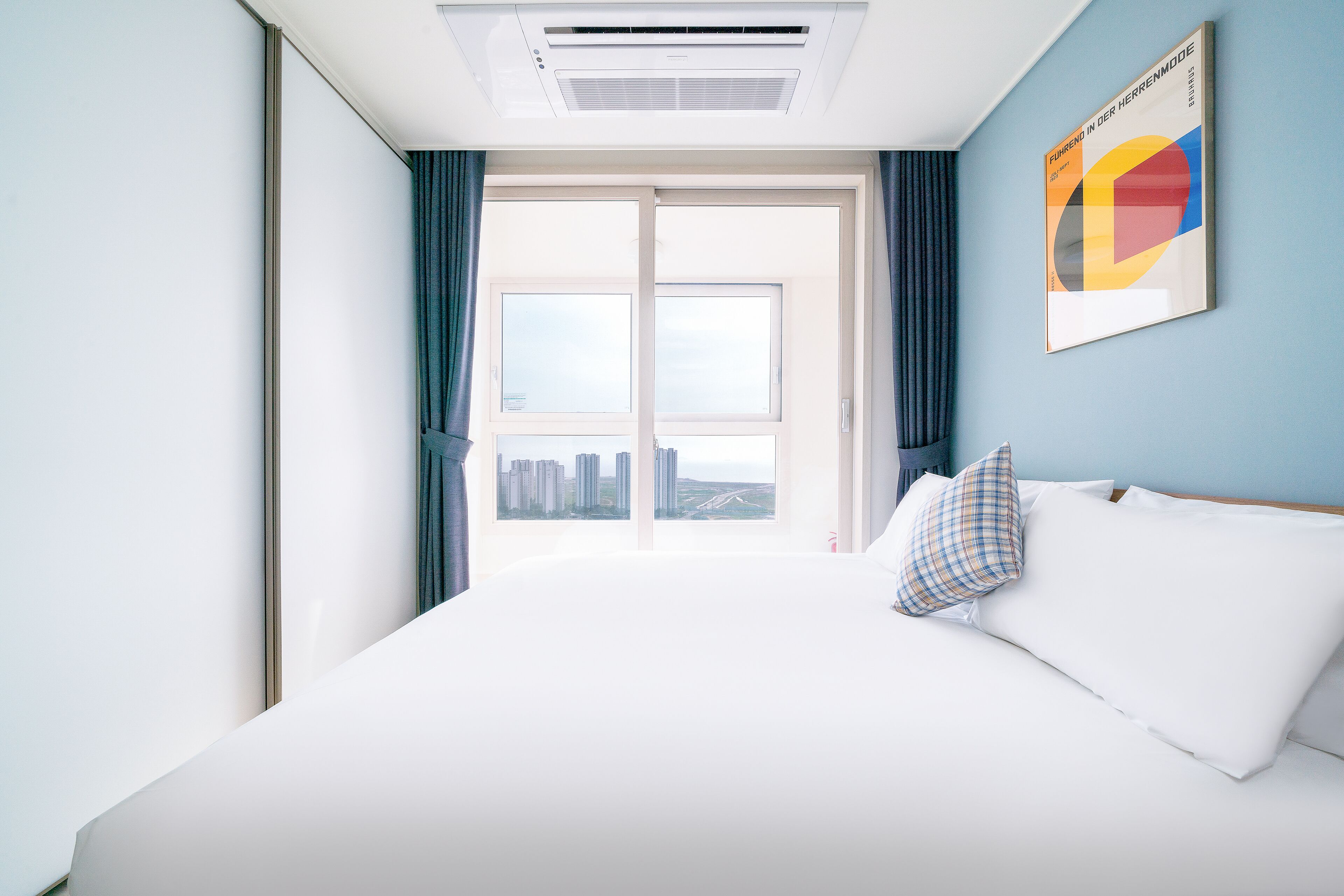 Habitación de hotel con una cama grande, vista de la ciudad a través de una puerta corredera y aire acondicionado en el techo.