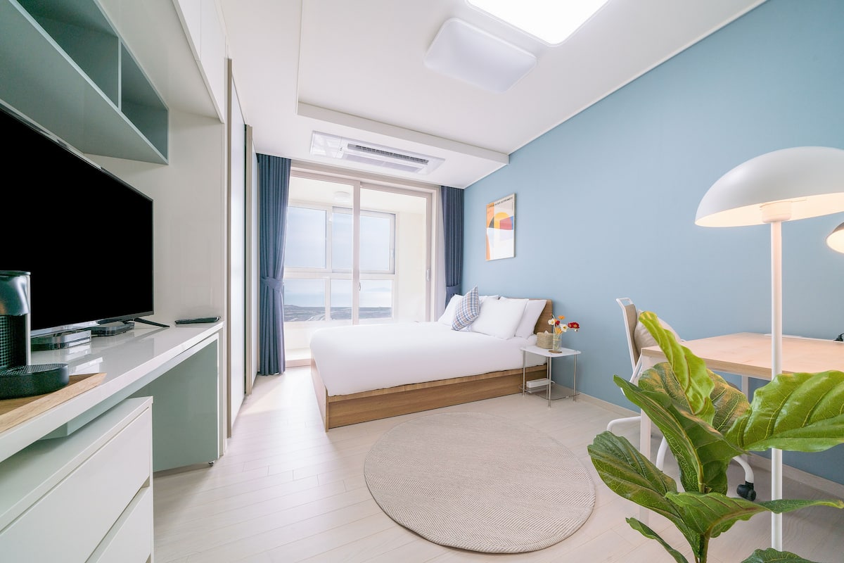 Una habitación de hotel moderna con cama, mesita de noche, lámpara y un ventanal grande.