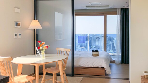 A modern hotel room with a bed, a dining area with a table and chairs, a lamp, and a view of the cityscape through large windows. - Urbanstay Jeju Airport (Jeju City)