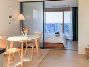 A modern hotel room with a bed, a dining area with a table and chairs, a lamp, and a view of the cityscape through large windows. - Urbanstay Jeju Airport (Jeju City)