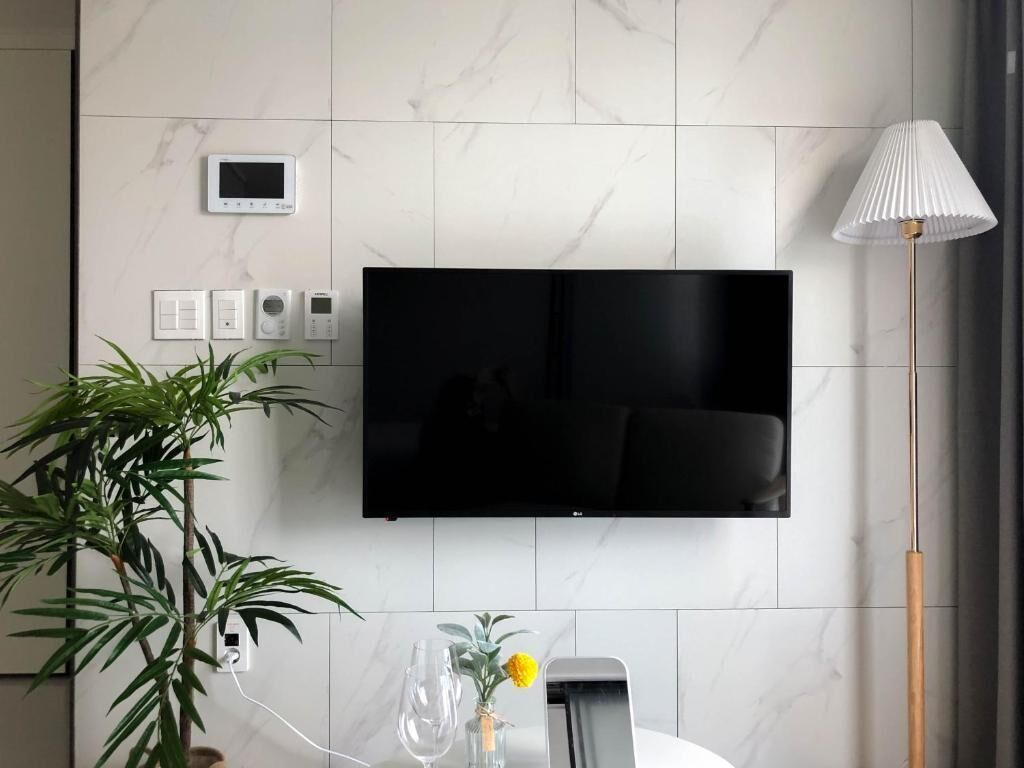 A modern living room with a flat-screen TV mounted on a marble wall, a potted plant, a vase with flowers, and a standing lamp.
