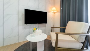 A modern hotel room with a flat-screen TV mounted on the wall, a white armchair, a small round table with a vase of flowers, and a floor lamp. - Urbanstay Jeju Airport (Jeju City)