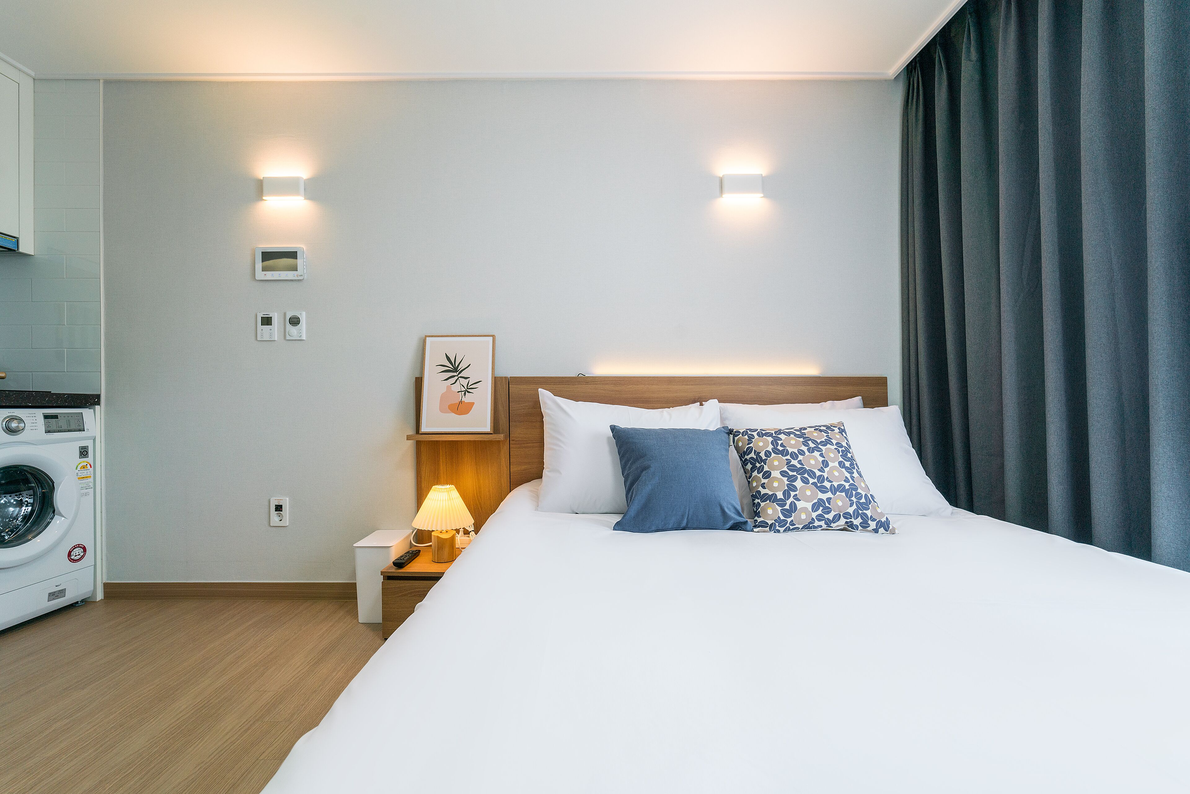 Hotelkamer met een bed, nachtkastje met lamp, wandbeugelthermostaat en een wasmachine in de hoek.