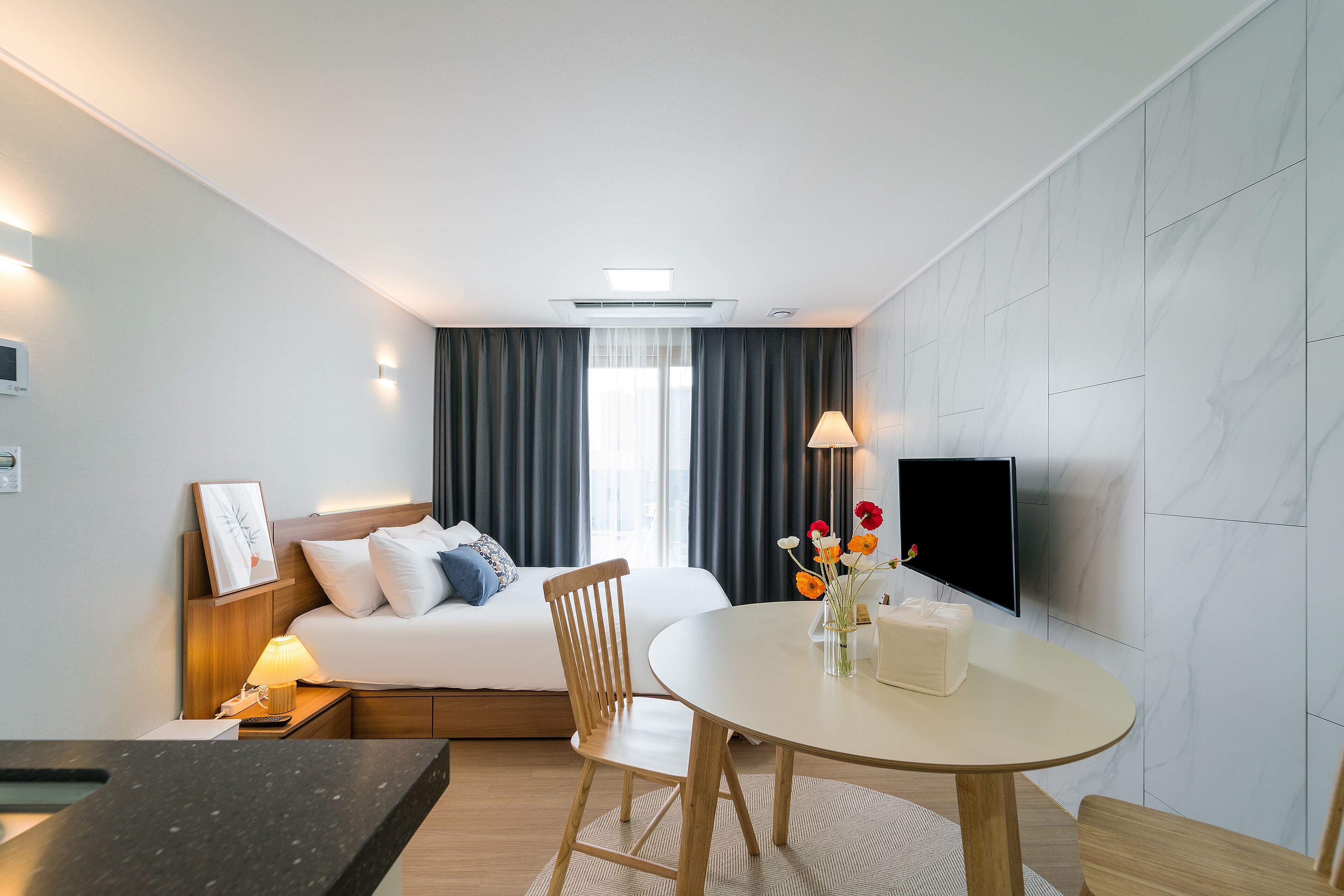 A hotel room with a bed, bedside table, lamp, TV, and a dining area with a table and chairs.